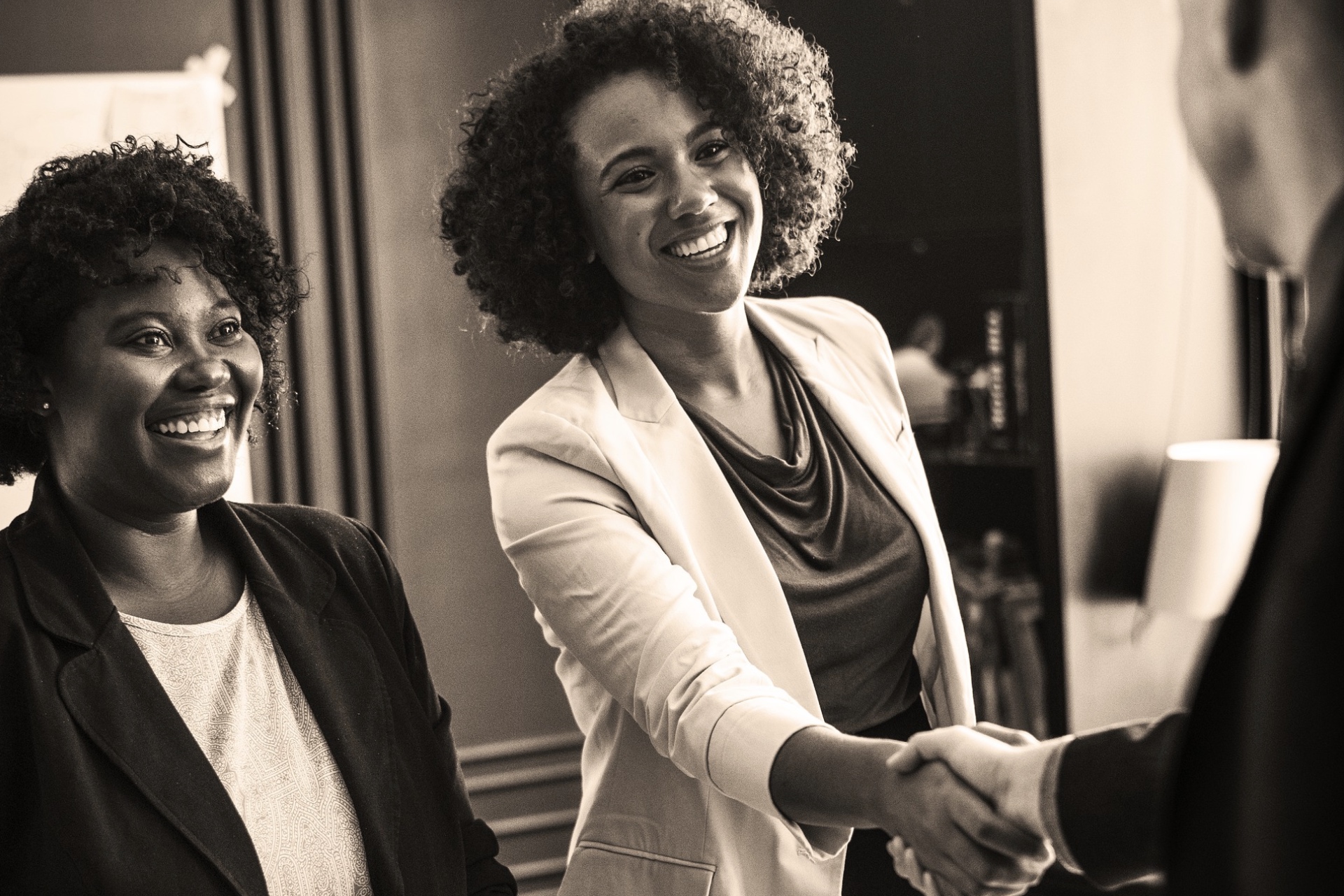 Black woman smiling, shaking hands with a man with another black woman standing next to her also smiling